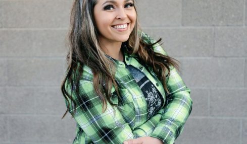 Smiling woman with long hair wearing a green plaid shirt standing against a gray brick wall.