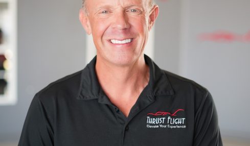 Smiling man in black polo shirt standing indoors with a blurred background and red text. Word 'FLIGHT' visible.