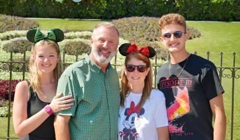 Four people smiling outdoors, two women and two men, with a green hedge and decorative elements in the background.
