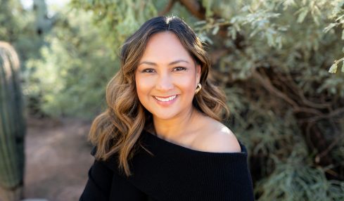 Woman with wavy hair smiling outdoors, wearing a black off-shoulder top, surrounded by greenery.