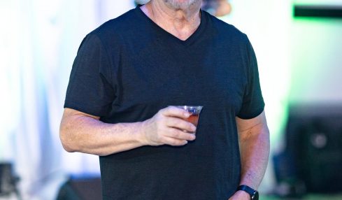 Man with glasses and a beard holding a drink, wearing a black t-shirt, at an indoor event with colorful lighting.