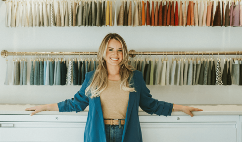 Woman standing in fabric store with colorful fabric swatches on wall behind her, smiling, arms outstretched, in a well-lit shop.